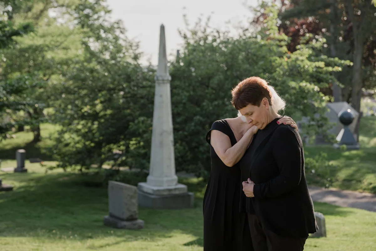 Two women in mourning embracing at a cemetery.